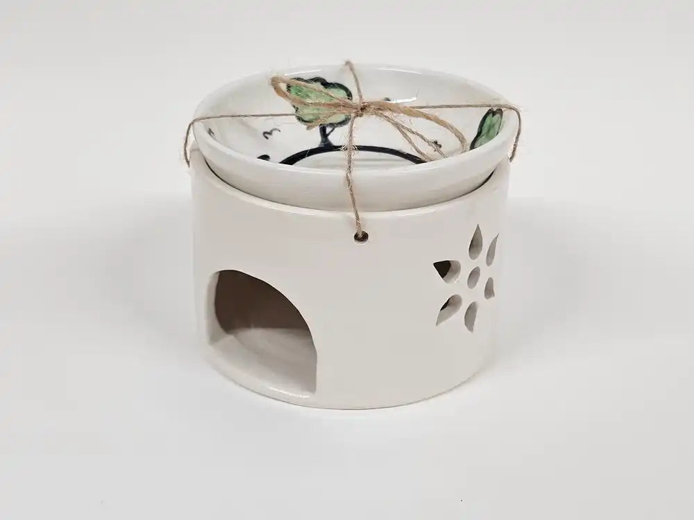 A white ceramic candle warmer and holder with a matching lid decorated with green and black floral motifs, tied with a piece of twine. The warmer features a cutout on the side and a small arched opening on the front.