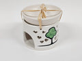 A white ceramic candle warmer with a matching lid, decorated with a green tree and black outline on the side, tied with a beige ribbon.
