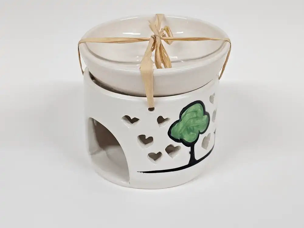 A white ceramic candle warmer with a matching lid, decorated with a green tree and black outline on the side, tied with a beige ribbon.