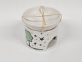 A white ceramic candle warmer with a matching lid, decorated with a green tree and black star and moon cutouts, tied with a natural twine bow.