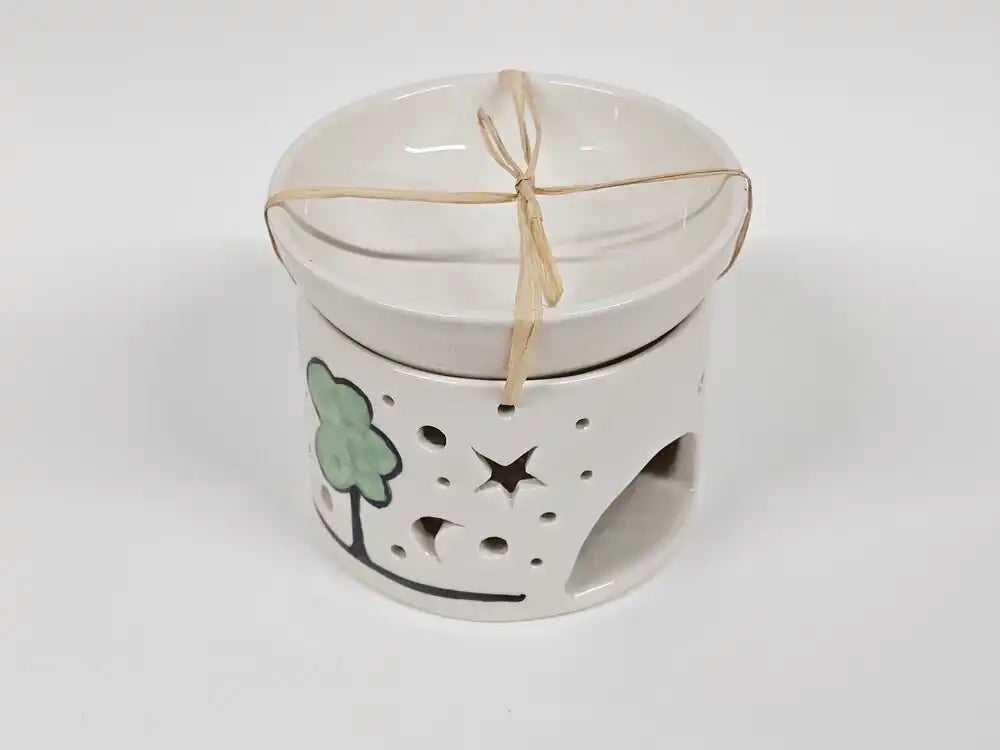 A white ceramic candle warmer with a matching lid, decorated with a green tree and black star and moon cutouts, tied with a natural twine bow.