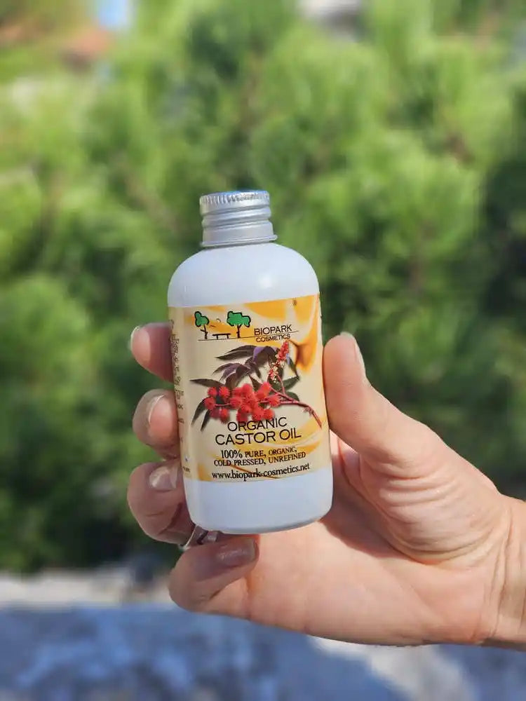 A hand holding a white bottle of Organic Castor Oil with a silver cap, set against a blurred green outdoor background.
