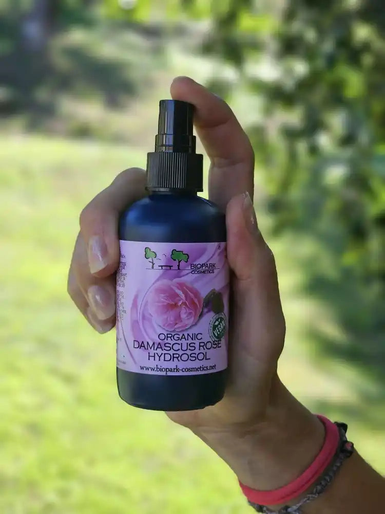 A hand holding a bottle of BioPark Cosmetics Organic Damascus Rose Hydrogel in front of a blurred green outdoor background.