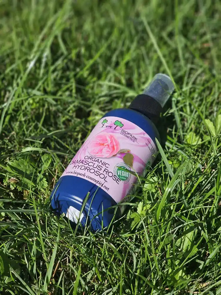 A bottle of BioMark Organic Amasculus Rose Hydrosol massage oil lies on green grass. The bottle is blue with a pink label featuring a rose graphic and a black spray nozzle.