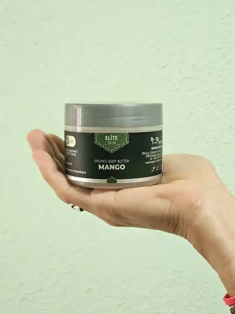 A hand holding a silver and black jar of Elite Skin Organic Body Butter in Mango against a light green textured wall.