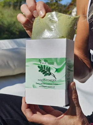 A person holds a white box labeled 'INDIGO POWDER' with a green leaf graphic, while pulling a sealed green sachet out of the top. The photo is taken outdoors with a blurred background.