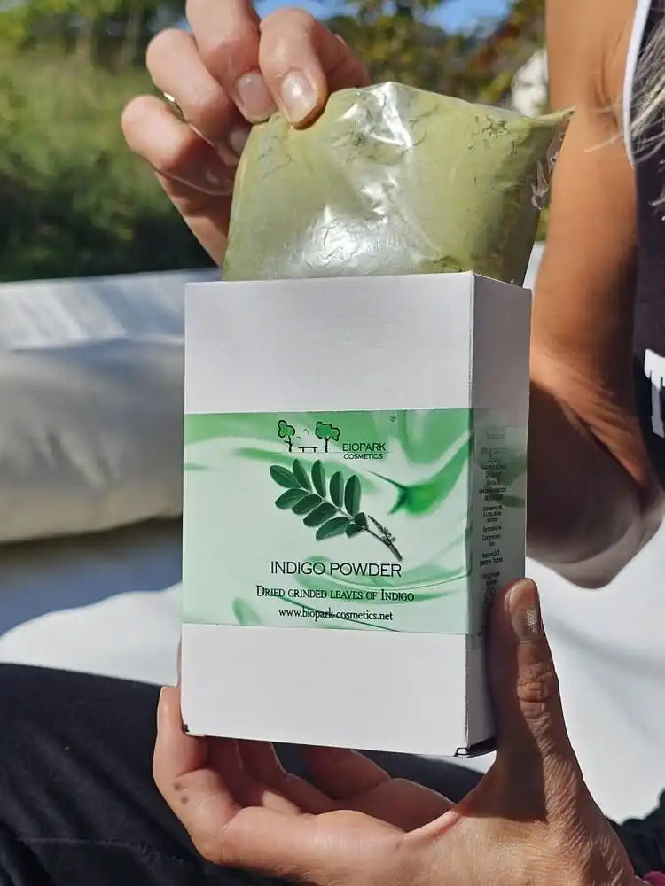 A person holds a white box labeled 'INDIGO POWDER' with a green leaf graphic, while pulling a sealed green sachet out of the top. The photo is taken outdoors with a blurred background.