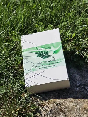 A white rectangular box of Indigo Powder (Dye Plant Leaves Extract) resting on a patch of green grass and stone outdoors.