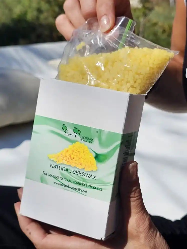 A person holds a white box labeled 'Natural Beeswax' with a clear plastic bag filled with yellow wax beads above the open box, photographed outdoors with a blurred light-colored surface and greenery in the background.