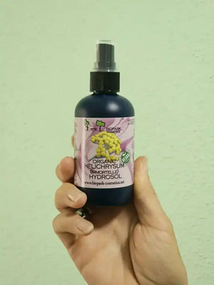 A hand holding a dark blue spray bottle of BioWick Organic Helichrysum (Borage) Hydrosol against a light green textured wall.