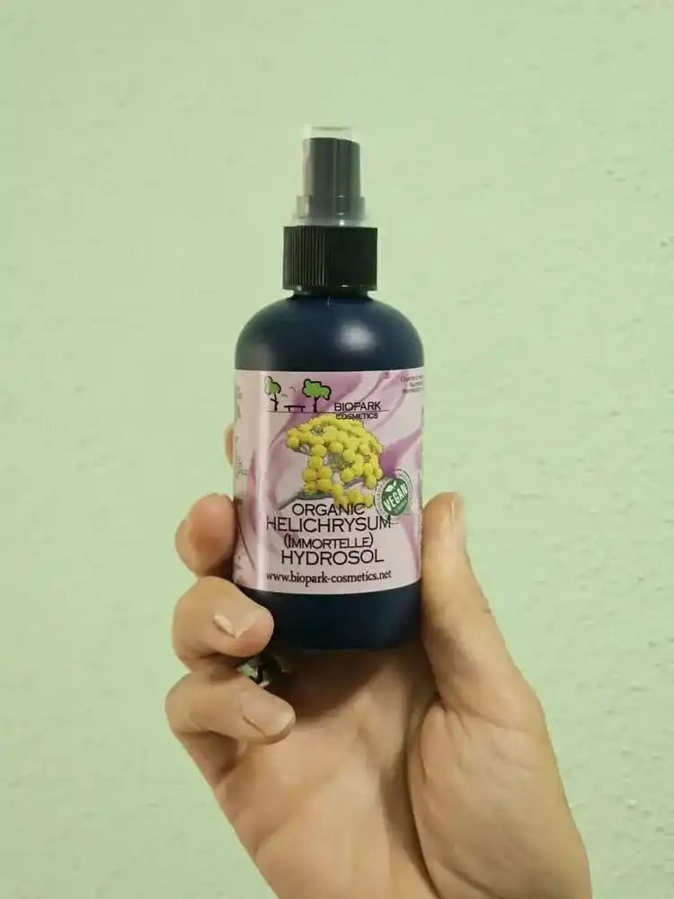 A hand holding a dark blue spray bottle of BioWick Organic Helichrysum (Borage) Hydrosol against a light green textured wall.