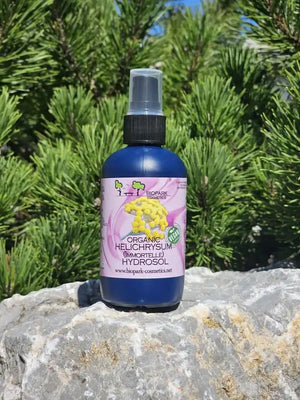 A bottle of Helichrysum Hydrosol facial mist by Naturpark Kalkafelden, placed on a gray stone with green pine trees in the background.