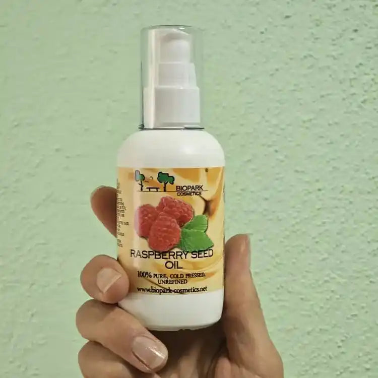 A hand holding a white pump bottle of BioMark Organics Raspberry Seed Oil against a textured light green wall.