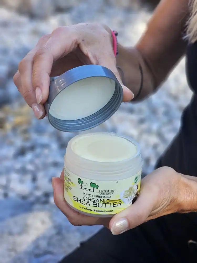 A person holds an open jar of Organic Shea Butter outdoors, with the lid held up to show the white balm inside. The jar has a white label with green and yellow graphics, and the person is wearing a black top and a pink bracelet.