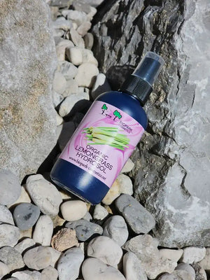 A bottle of Organic Lemongrass Hydrosol  lies on a bed of small gray and white pebbles, nestled between large rocks. The bottle is dark blue with a pink and white label, and it has a black spray nozzle.